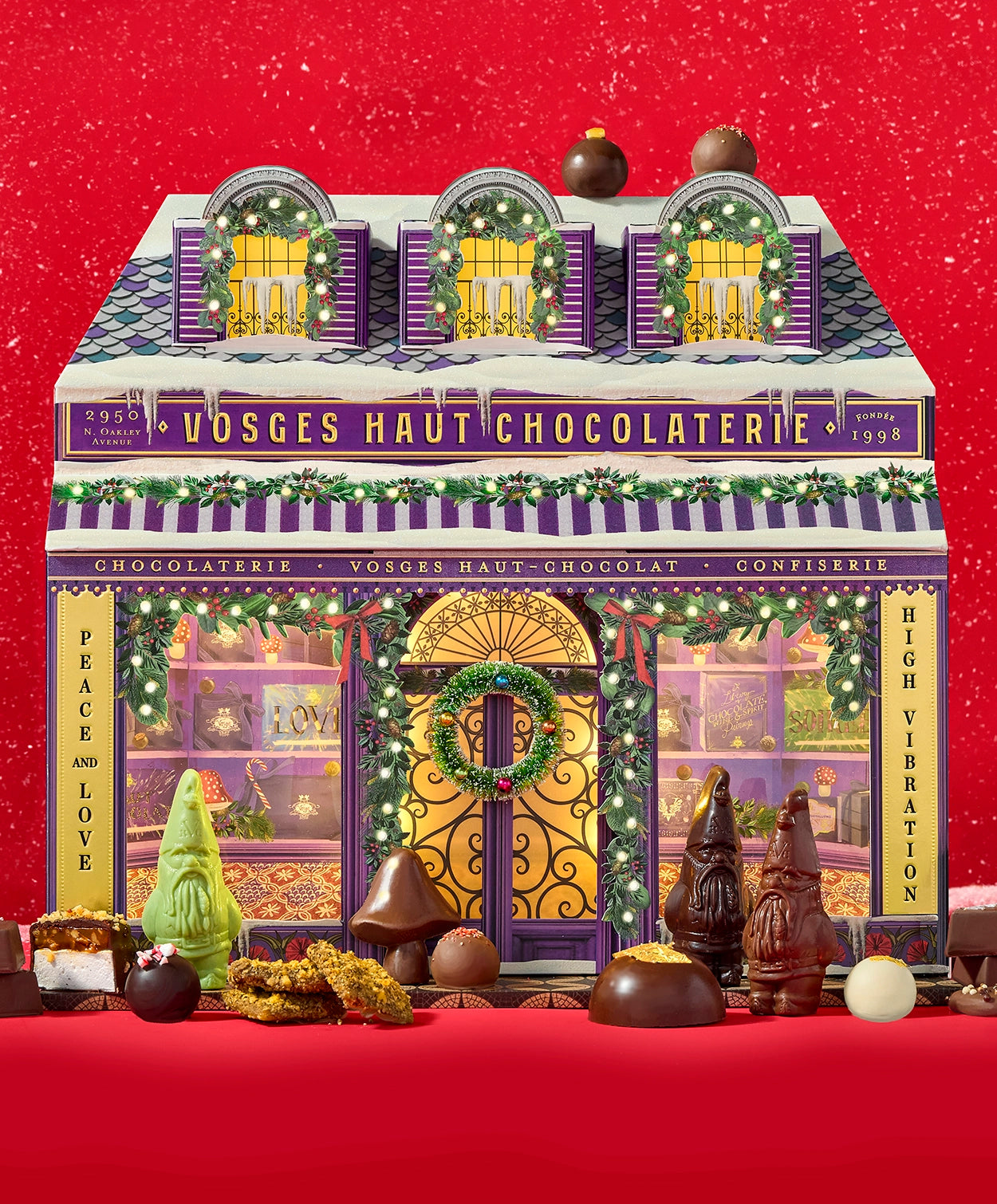 A Chocolate Advent calendar resembling a small chocolate shop sits on a red background surround by chocolate confections and lightly falling snow.