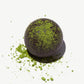 Close up view of a Vosges Black Pearl truffle topped with bright green matcha powder on a light grey background.