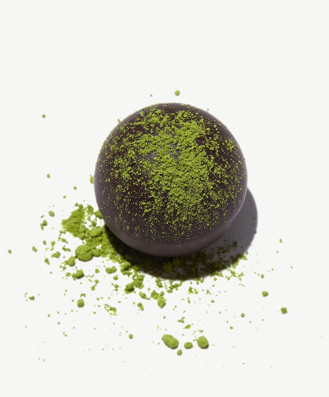 Close up view of a Vosges Black Pearl truffle topped with bright green matcha powder on a light grey background.