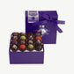 A purple candy box tied with a purple ribbon bow sits upright displaying sixteen dark chocolate truffles adorned in colorful toppings and chopped nuts on a light grey background.
