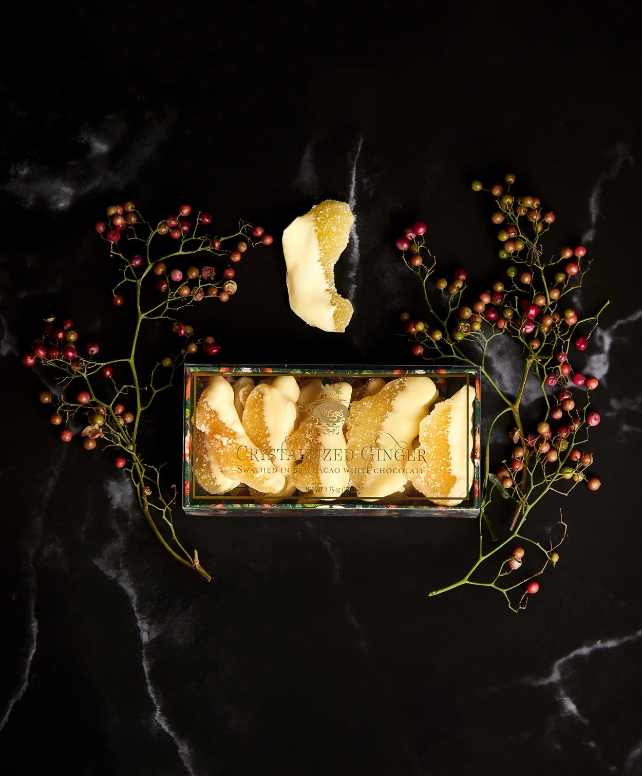 A piece of Vosges dipped crystalized ginger beside a branch of dried berries and a box of ginger on a black marble background.