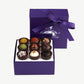 Purple box of Vosges haut-chocolat exotic truffles, lid standing upright, displaying nine chocolate truffles adorned in brightly colored spices and toppings tied with a purple ribbon bow on a white background.