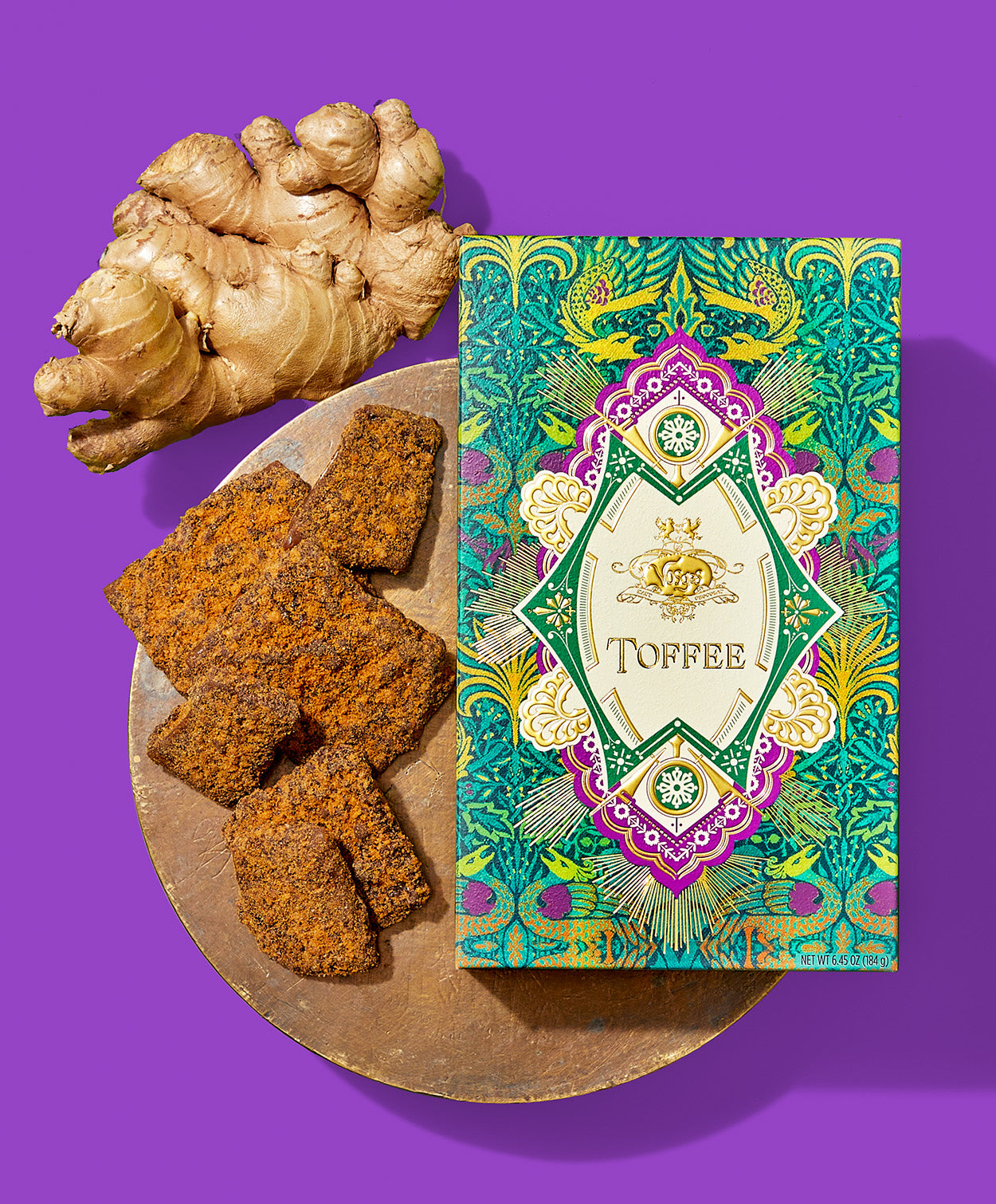 A green and purple gable box sits beside several pieces of Gingerbread Toffee on a wooden tray beside a large ginger root on a bright purple background.