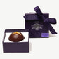 A large hazelnut Reishi mushroom chocolate bombalina truffle in an opened purple candy box standing upright tied with a purple ribbon bow on a white background.