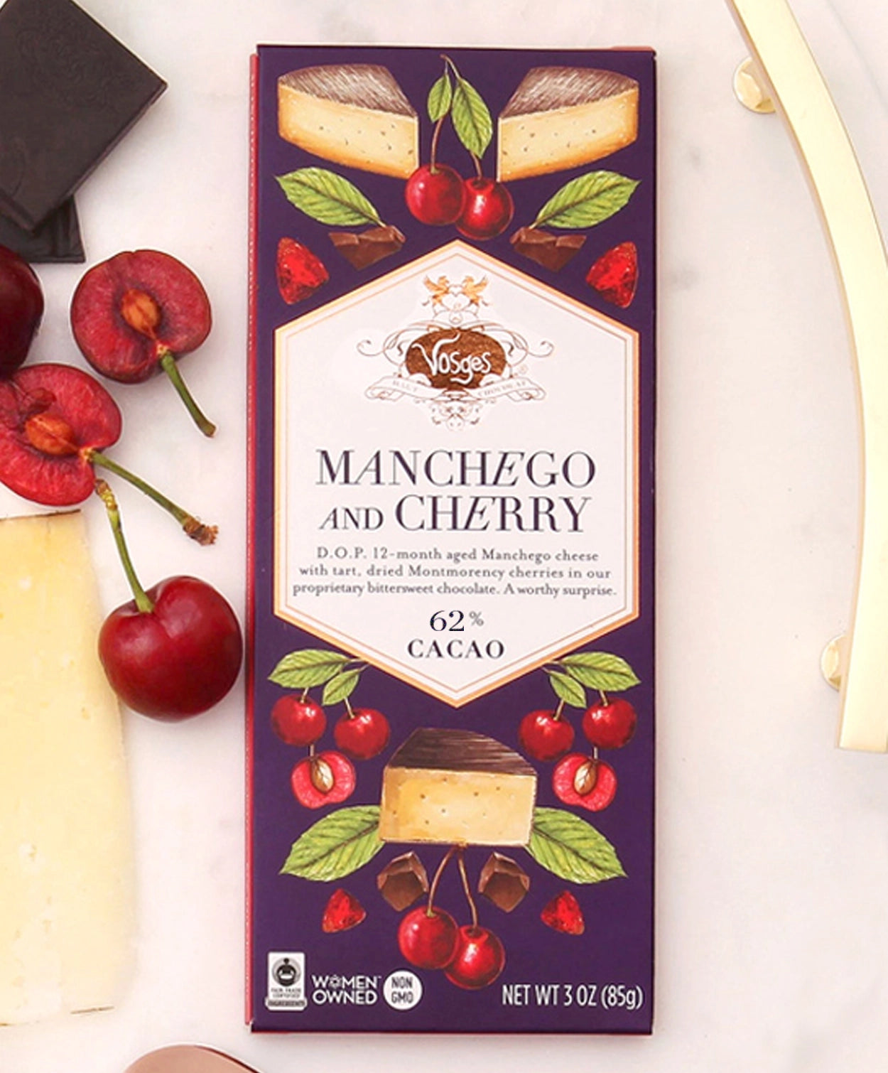 A 3 ounce Vosges Manchego and Cherry bar beside cheese a few cherries and pieces of chocolate on a white background