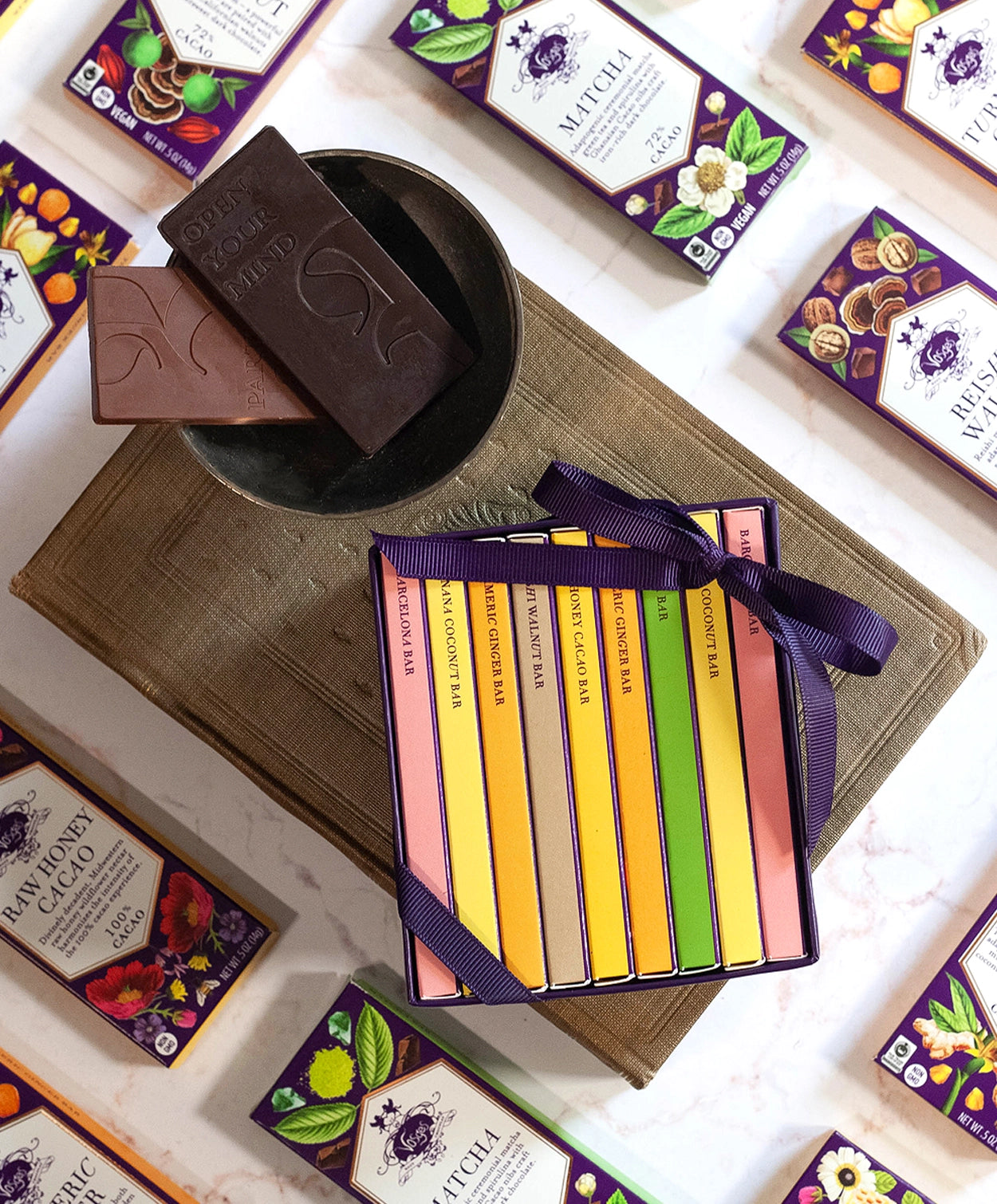 Vosges Mini Chocolate Bar Library and two bars of chocolate sit on an antique book surrounded by mini chocolate bars on a marble table top.