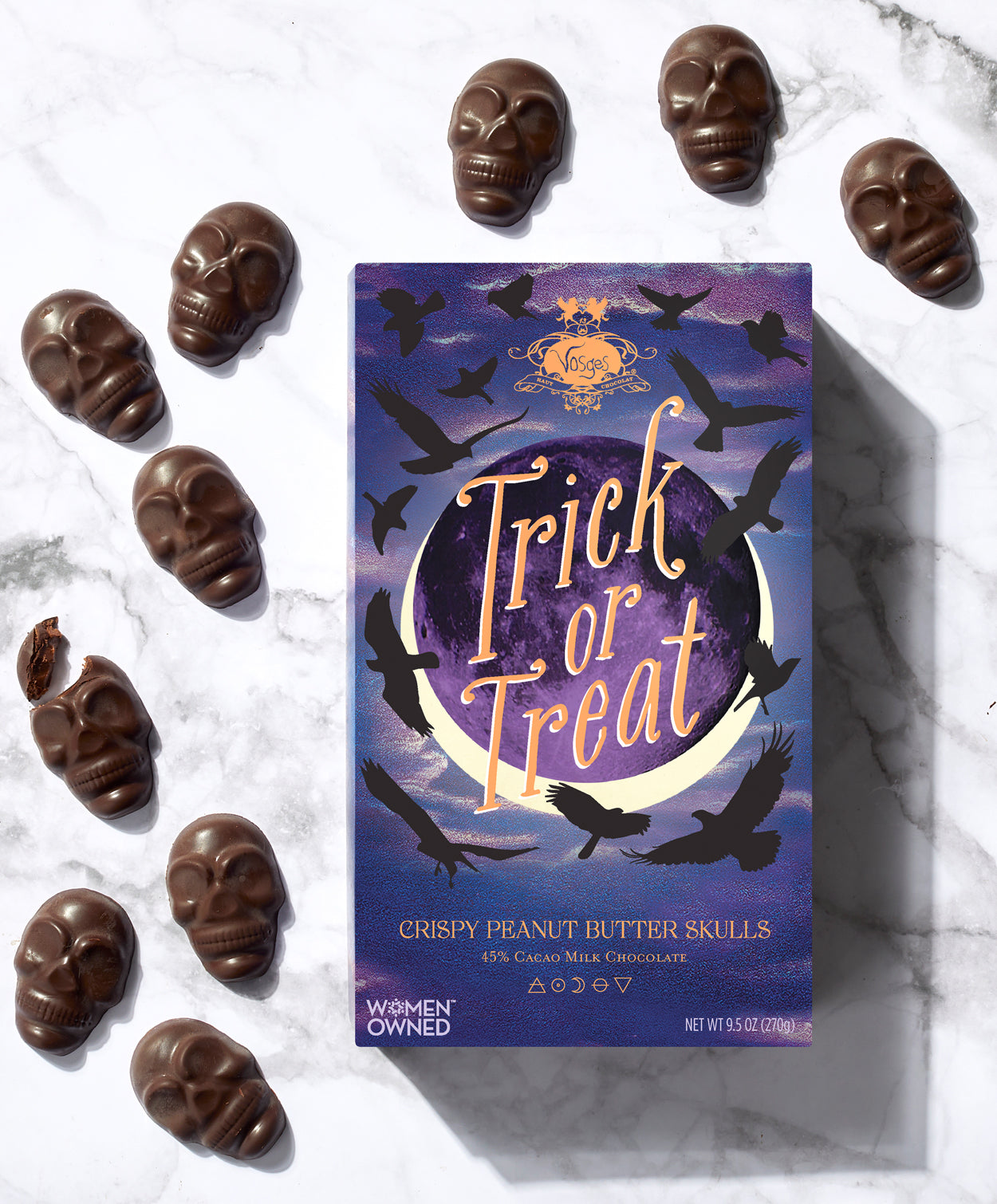 A purple and blue box reading, "Trick or Treat" along with several chocolate skulls sits on a marble background.