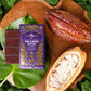 Vosges Raw Almond Butter Bar on a large green leaf beside a cacao pod surrounded by green moss. 