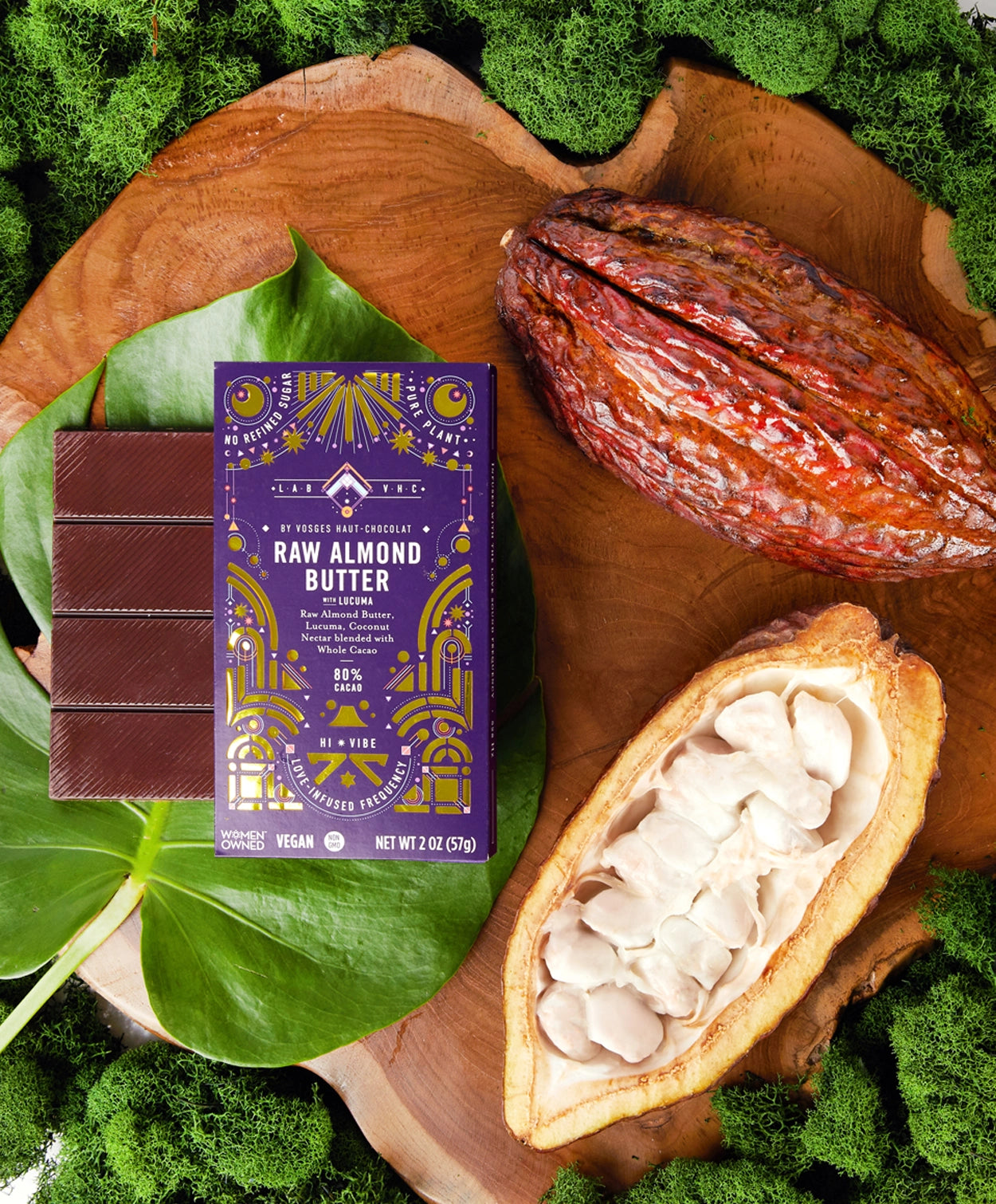 Vosges Raw Almond Butter Bar on a large green leaf beside a cacao pod surrounded by green moss. 