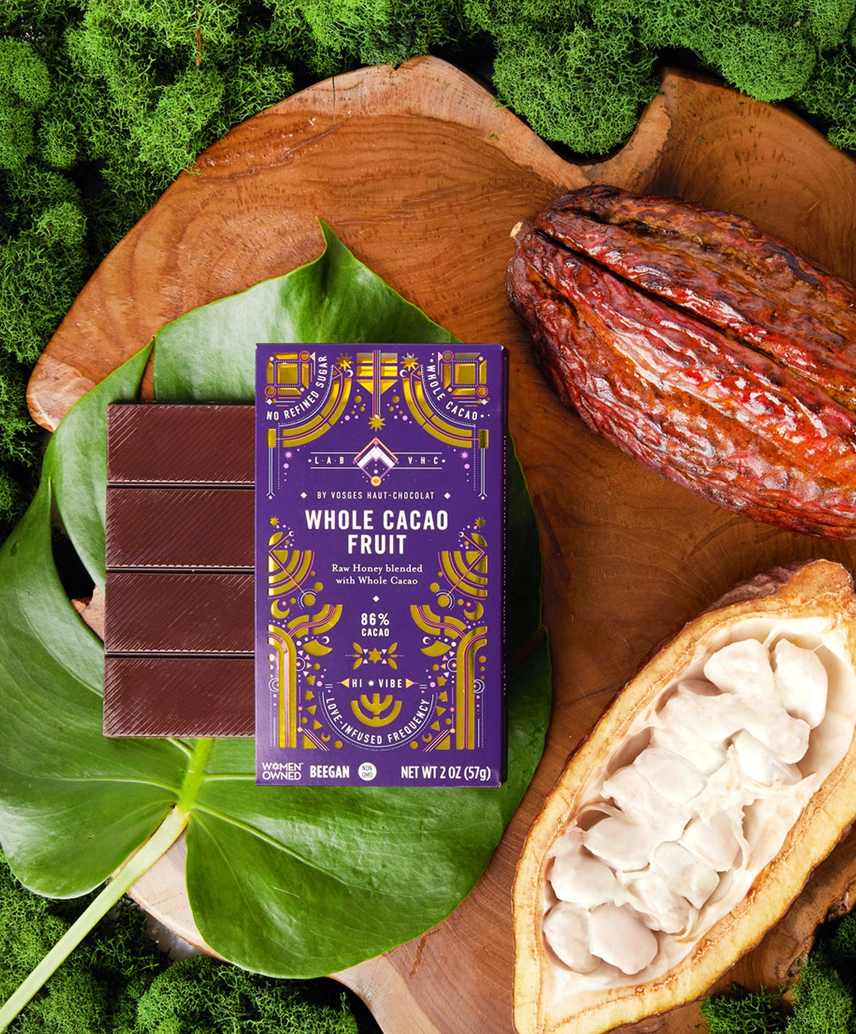 Vosges Whole Cacao Fruit Pure Plant Bar on a large green leaf beside an opened cacao pod surrounded by green moss.