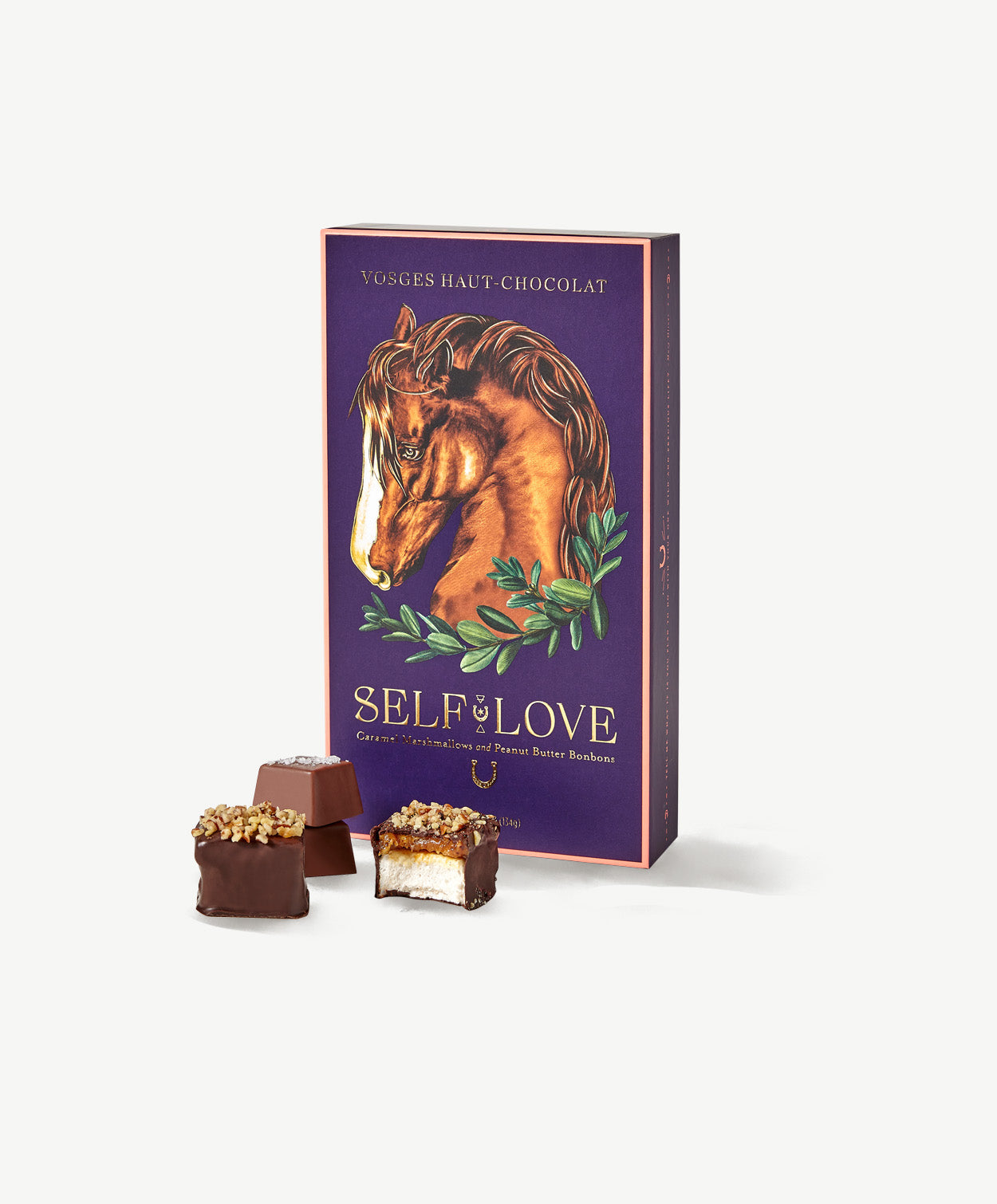 A tall purple candy box decorated with a hand-drawn horse sits behind three stacks of Vosges caramel marshmallows and peanut butter bonbons. on a grey background.