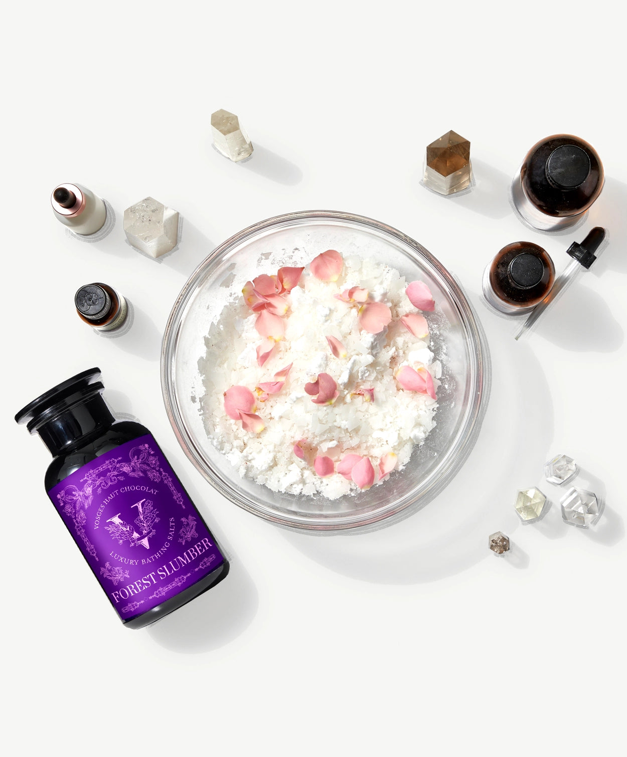 Vosges Haut Chocolat Forest Slumber bath salts beside several crystals and a bowl of flower petals on a light grey background.