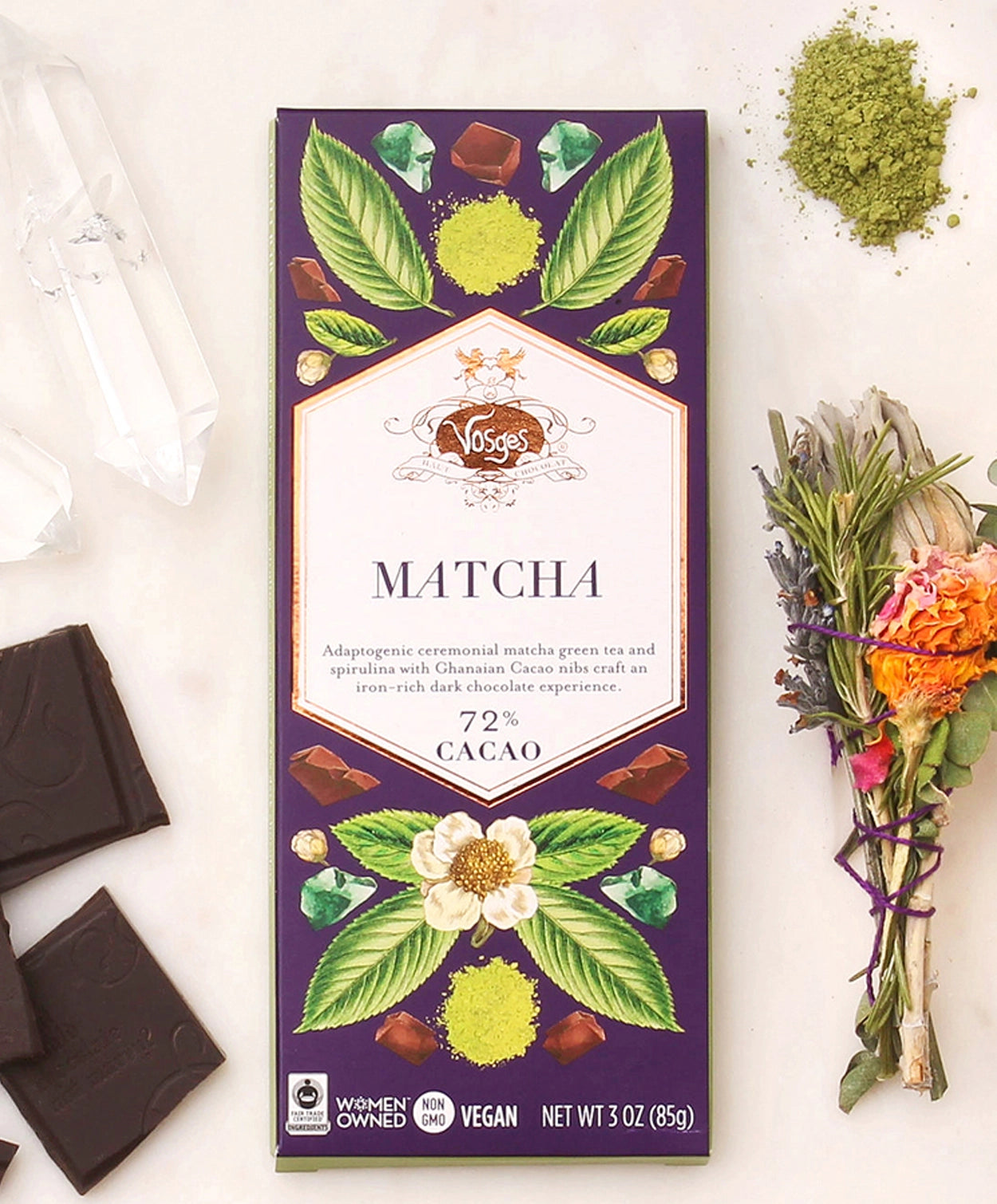 Vosges Matcha Green Tea Spirulina chocolate bar beside a bundle of dried flowers, green matcha powder, and several crystals on a white background.