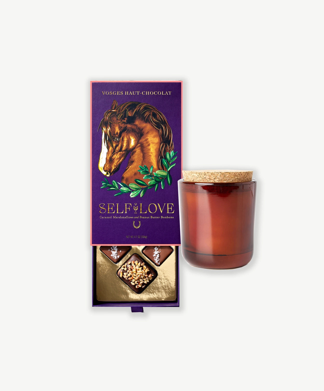 An opened Vosges chocolate box ornately decorated with a horse and gold foil sits open revealing peanut butter bon bons and a chocolate covered marshmallow beside a candle in a brown jar on a light grey background.