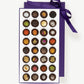 Large purple candy box of Vosges Haut-Chocolat sits open displaying thirty two chocolate truffles adorned with colorful spices, chopped nuts and dried flowers tied with a purple ribbon bow on a grey background.