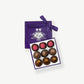 A small purple candy box tied with a purple ribbon bow sit's open displaying three rows of vegan chocolate truffles adorned in colorful toppings on a light grey background.
