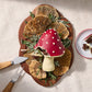 Red mushroom shaped chocolate dessert with white speckles on a wooden board surrounded by herbs and a knife