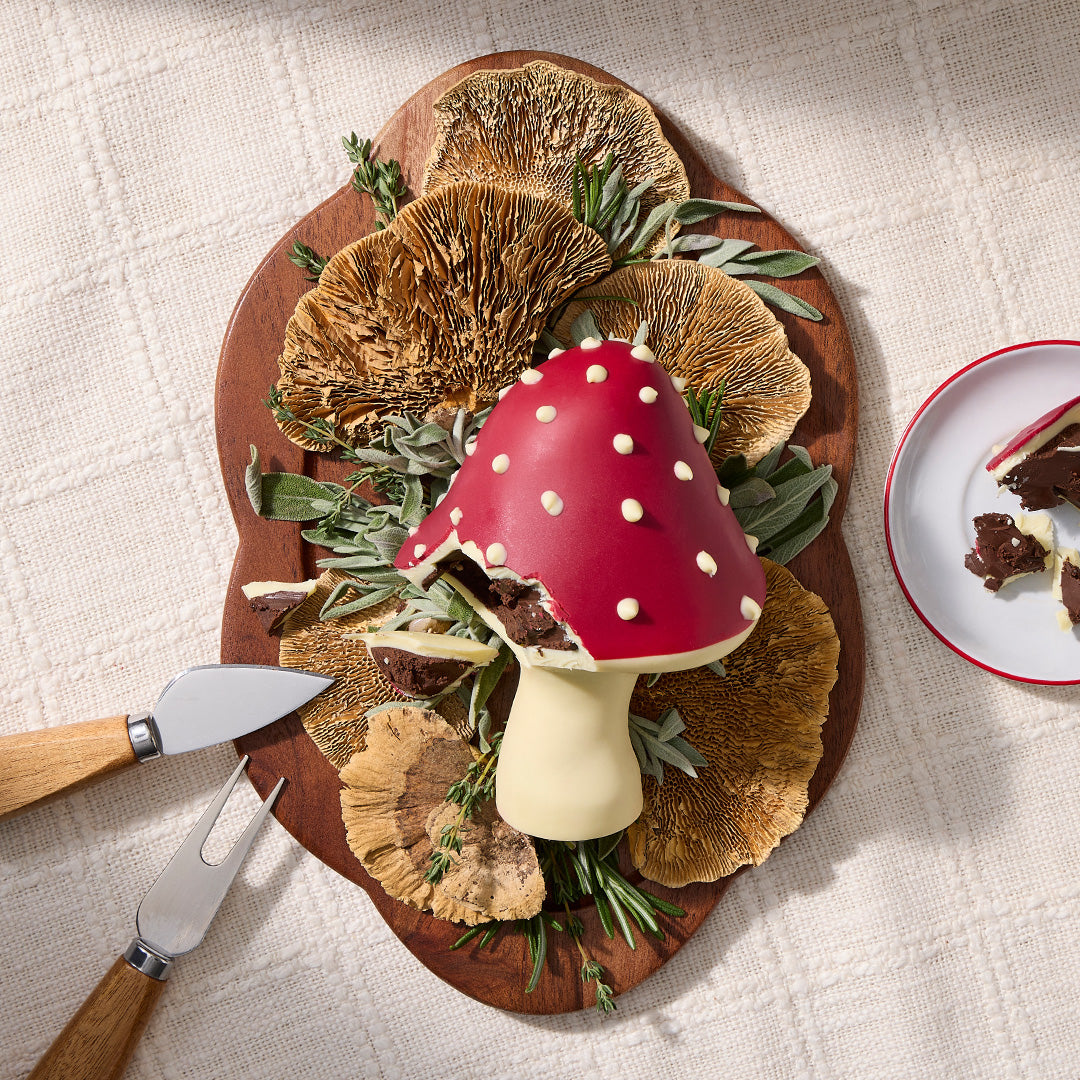 Red mushroom shaped chocolate dessert with white speckles on a wooden board surrounded by herbs and a knife