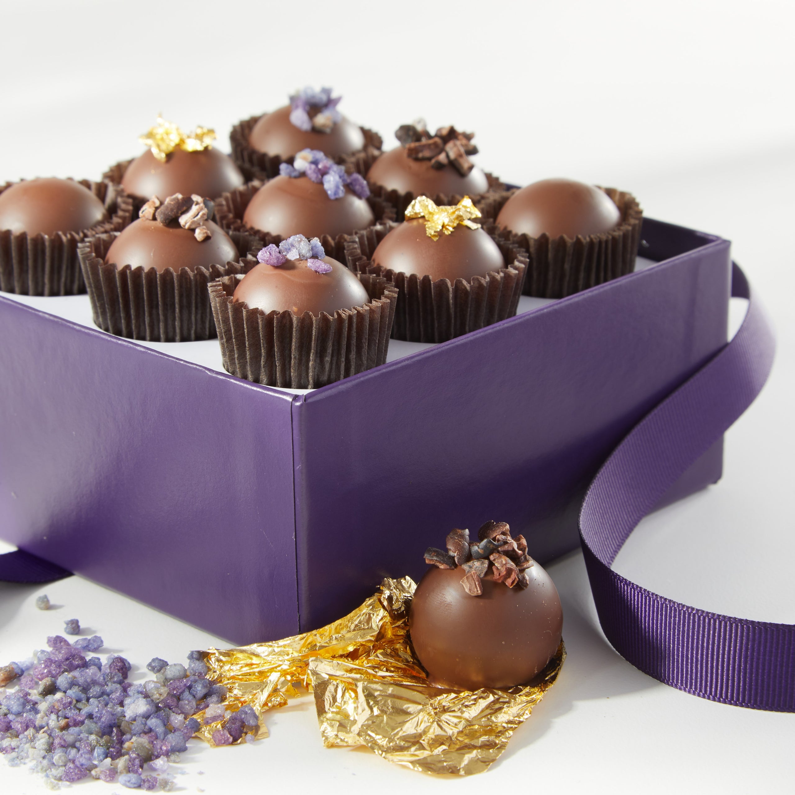Close up view of a Vosges Praline collection box opened revealing 9 milk chocolate truffles adorned with crystalized violets, coco nibs and 24kt gold leaf on a white background.