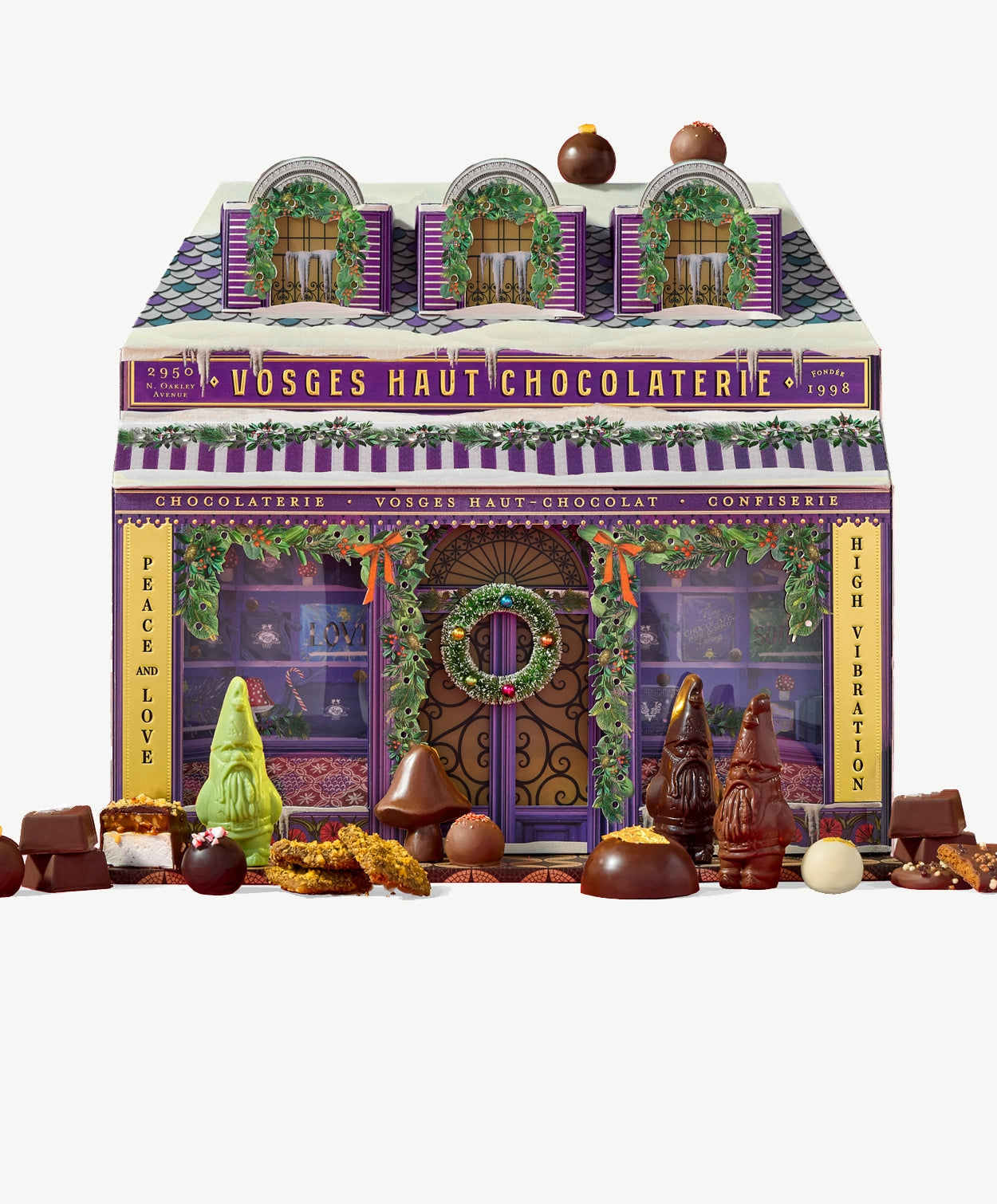 A Vosges Calendar of Advent resembling a small Chocolaterie, decorated in purple and gold foil sits surrounded by chocolate confections on a light grey background.