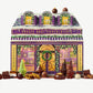 A Vosges Chocolate advent calendar decorated to resemble a small chocolaterie, adorned in christmas lights and greens sits surrounded by chocolate bonbons and gnomes on a light grey background.