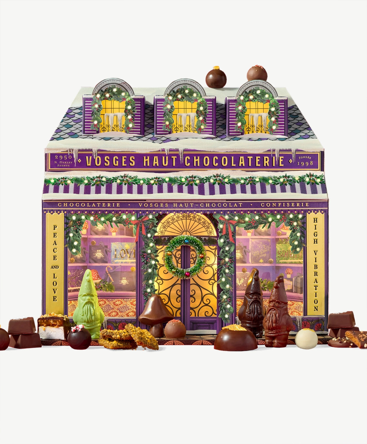 A Vosges Chocolate advent calendar decorated to resemble a small chocolaterie, adorned in christmas lights and greens sits surrounded by chocolate bonbons and gnomes on a light grey background.