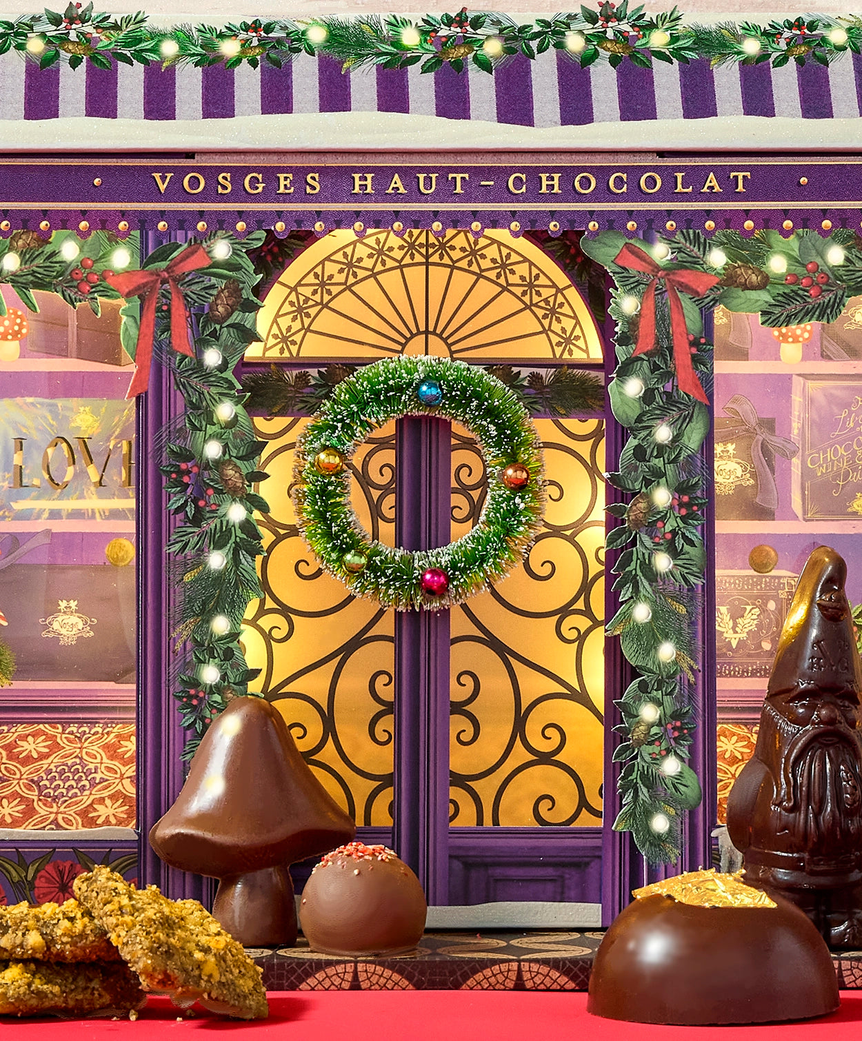 A closeup view of the Vosges Advent calendar: a miniature chocolate shop doorway entrance decorated with bright christmas lights, holly and a wreath surrounded by chocolate confections.