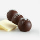 Three brown butter truffles with glossy chocolate coating topped with white flakes and a white chocolate piece nearby