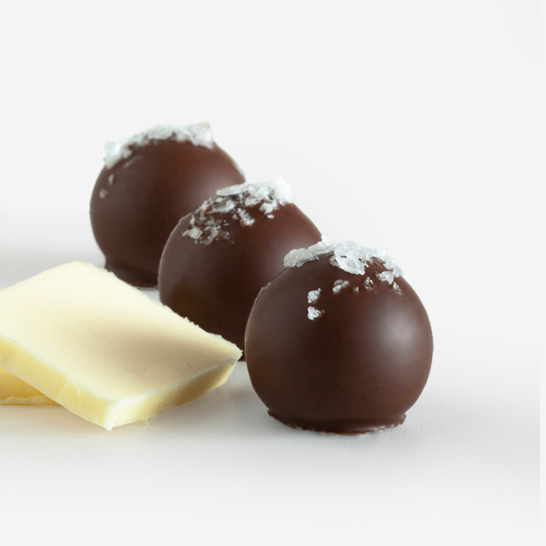 Three brown butter truffles with glossy chocolate coating topped with white flakes and a white chocolate piece nearby