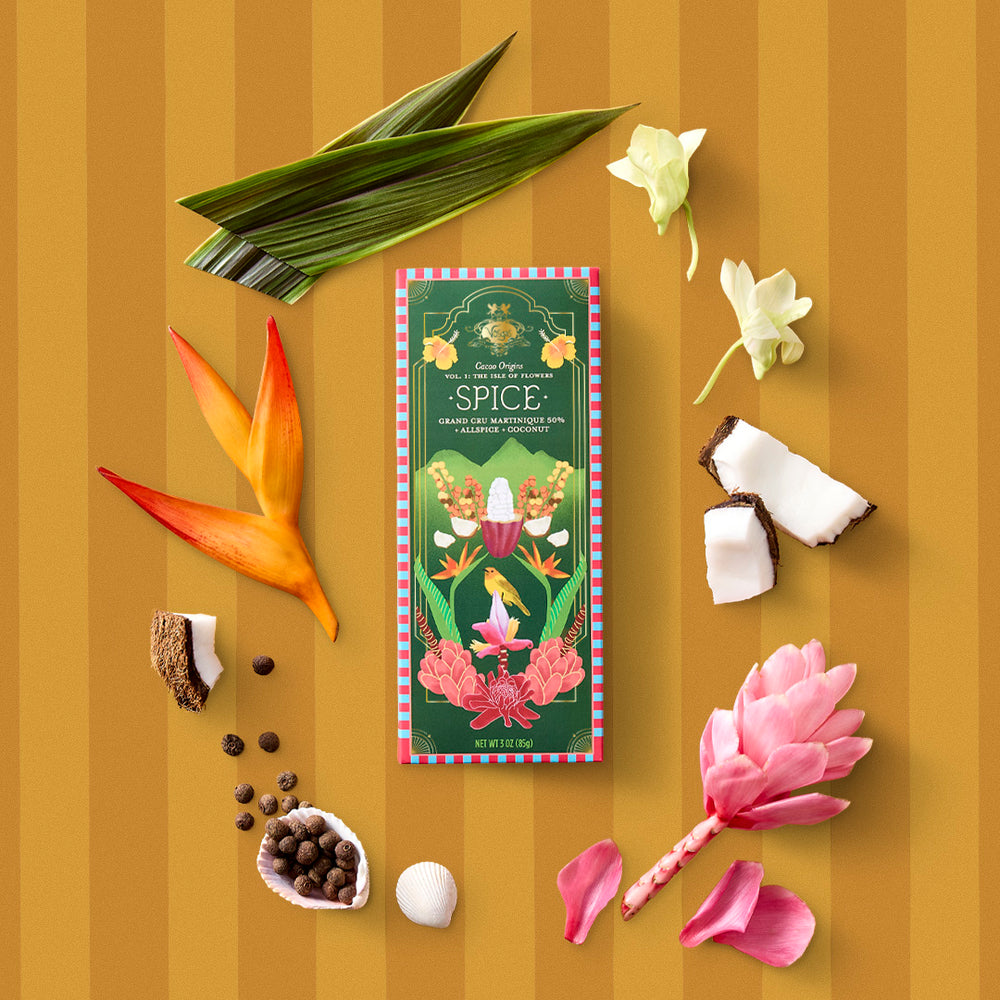 Martinique cacao single origin chocolate bar centered among tropical leaves blossoms and coconut pieces on a gold striped backdrop