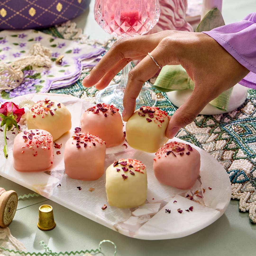 Hand reaching for a pink chocolate truffle on a decorative table with jewelry and a teacup.