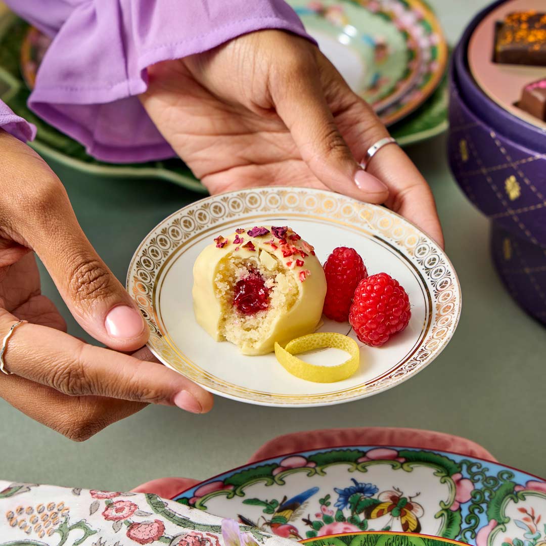 Person holding a small plate with a dessert and raspberries on a table with colorful plates.