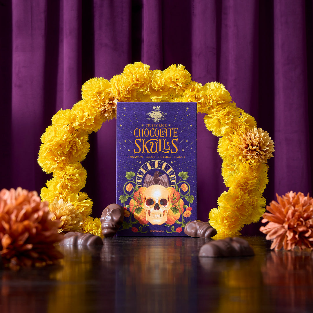 A boxed product labeled 'Chocolate Skulls' stands upright on a glossy dark surface, framed by a yellow flower garland and surrounded by chocolate pieces, against a purple curtain background.