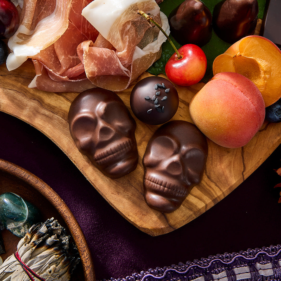 Two chocolate skull candies and one chocolate truffle with black seed details, arranged on a wooden board with prosciutto, cherries, apricots, and other food items.