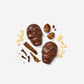 Two chocolate skull-shaped confections on a light gray background, surrounded by decorative ingredients including cinnamon sticks, cloves, and small pale yellow pieces. One skull is whole and the other is broken open to show the textured filling.