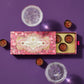 Top down view of a pink and gold Vosges chocolate box partially opened revealing several chocolate champagne dessert cups beside two glasses of bubbly wine on a light purple background.