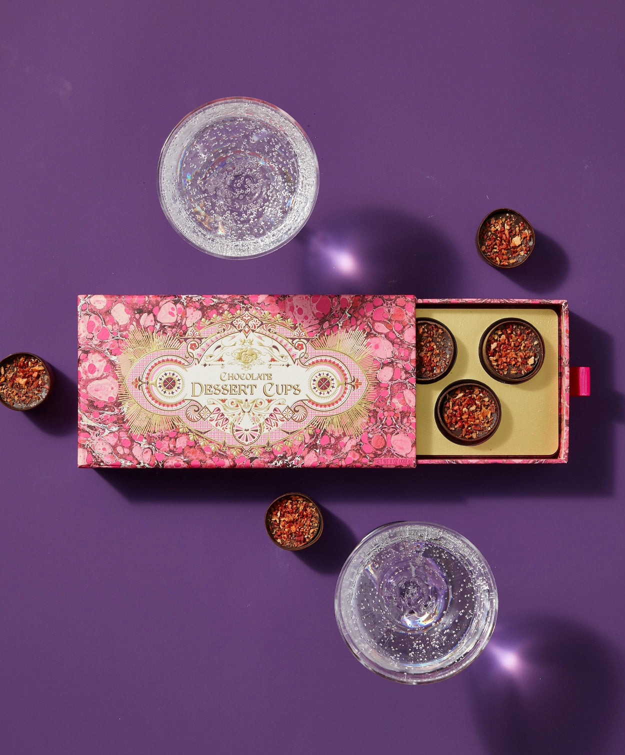 Top down view of a pink and gold Vosges chocolate box partially opened revealing several chocolate champagne dessert cups beside two glasses of bubbly wine on a light purple background.