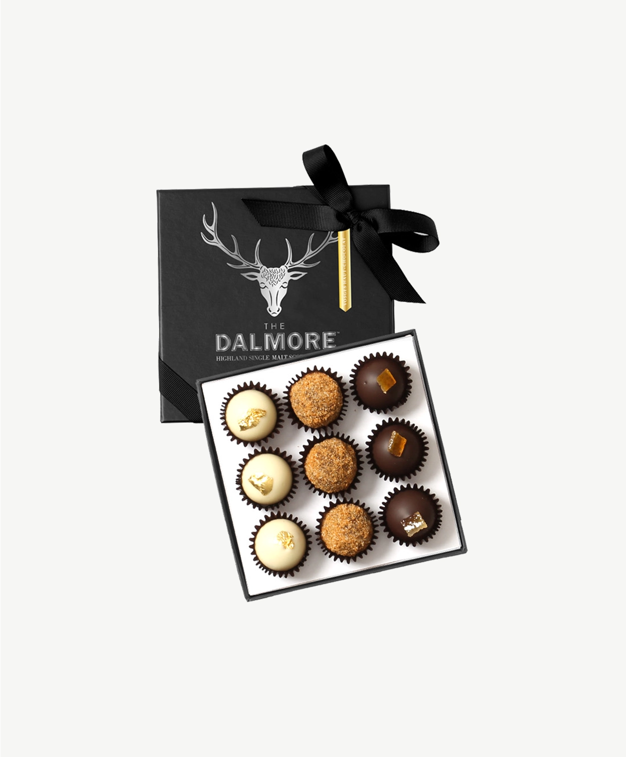 An open, black candy box adorned in silver foil tied with a black ribbon bow displays three rows of Vosges Dalmore chocolate truffles topped with spiced ginger crumb, gold leaf and candied orange peel sit in on a light grey background.