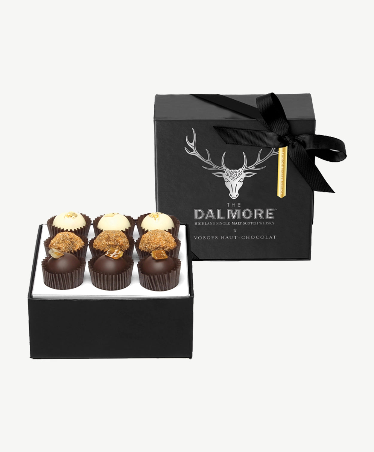 Nine Vosges Dalmore chocolate truffles topped with spiced ginger crumble, gold leaf and candied orange peel sit in a open, black candy box adorned in silver foil tied with a black ribbon bow on a white background.