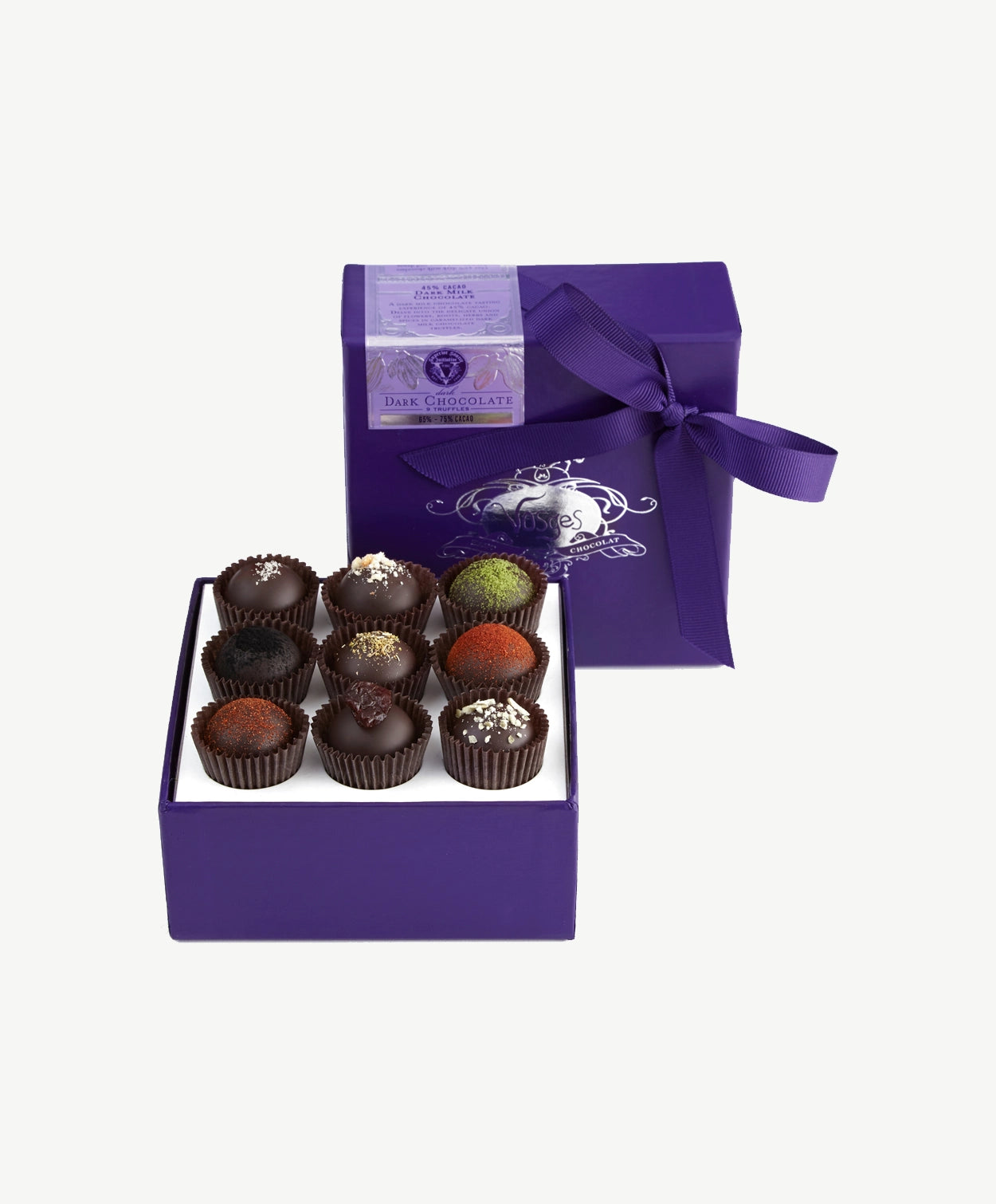 A small purple candy box tied with a purple ribbon bow sits upright displaying three rows of dark chocolate truffles adorned in colorful toppings and chopped nuts on a light grey background.