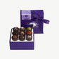 A small purple candy box tied with a purple ribbon bow sits upright displaying three rows of dark chocolate truffles adorned in colorful toppings and chopped nuts on a light grey background.