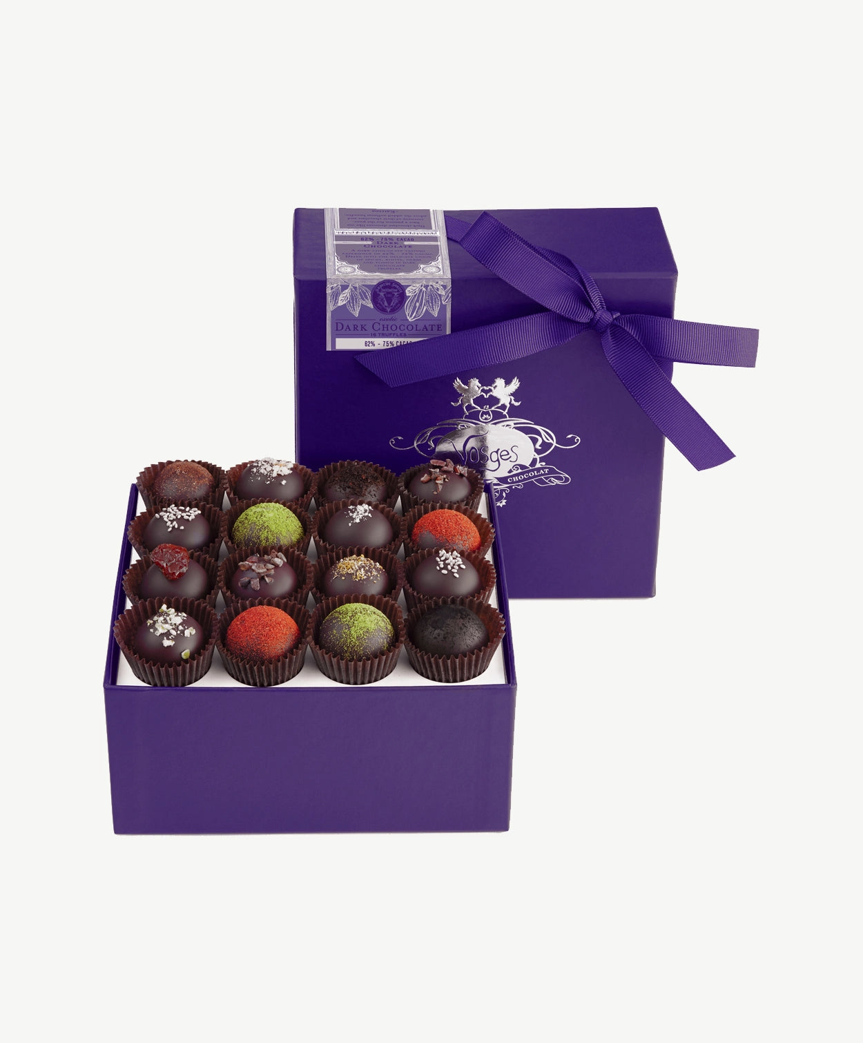 A purple candy box tied with a purple ribbon bow sits upright displaying sixteen dark chocolate truffles adorned in colorful toppings and chopped nuts on a light grey background.