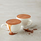 A pair of white espresso cups sit on a granite countertop brimming with foamy, Vosges La Parisienne  drinking chocolate.