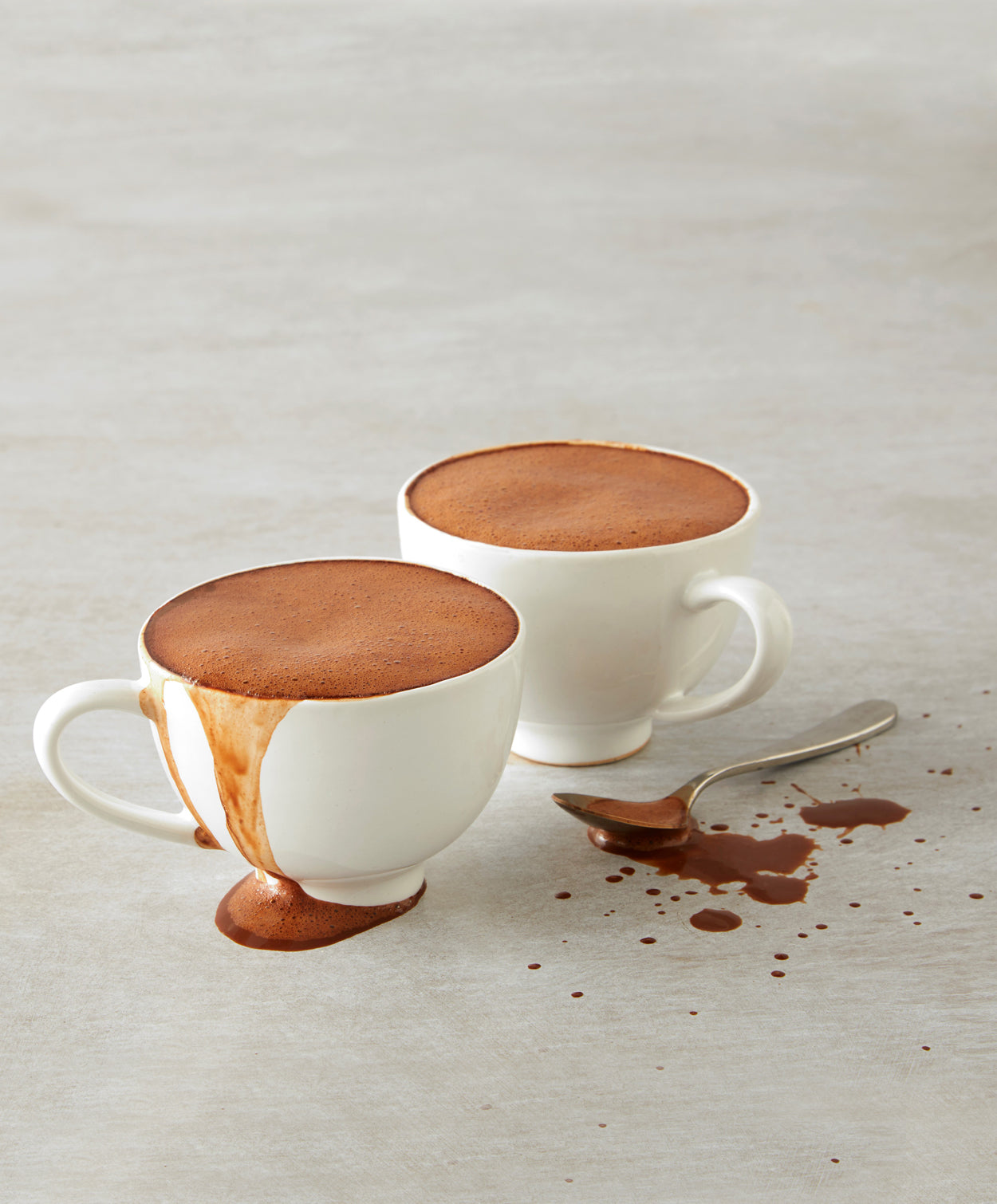 A pair of white espresso cups sit on a granite countertop brimming with foamy, Vosges La Parisienne  drinking chocolate.