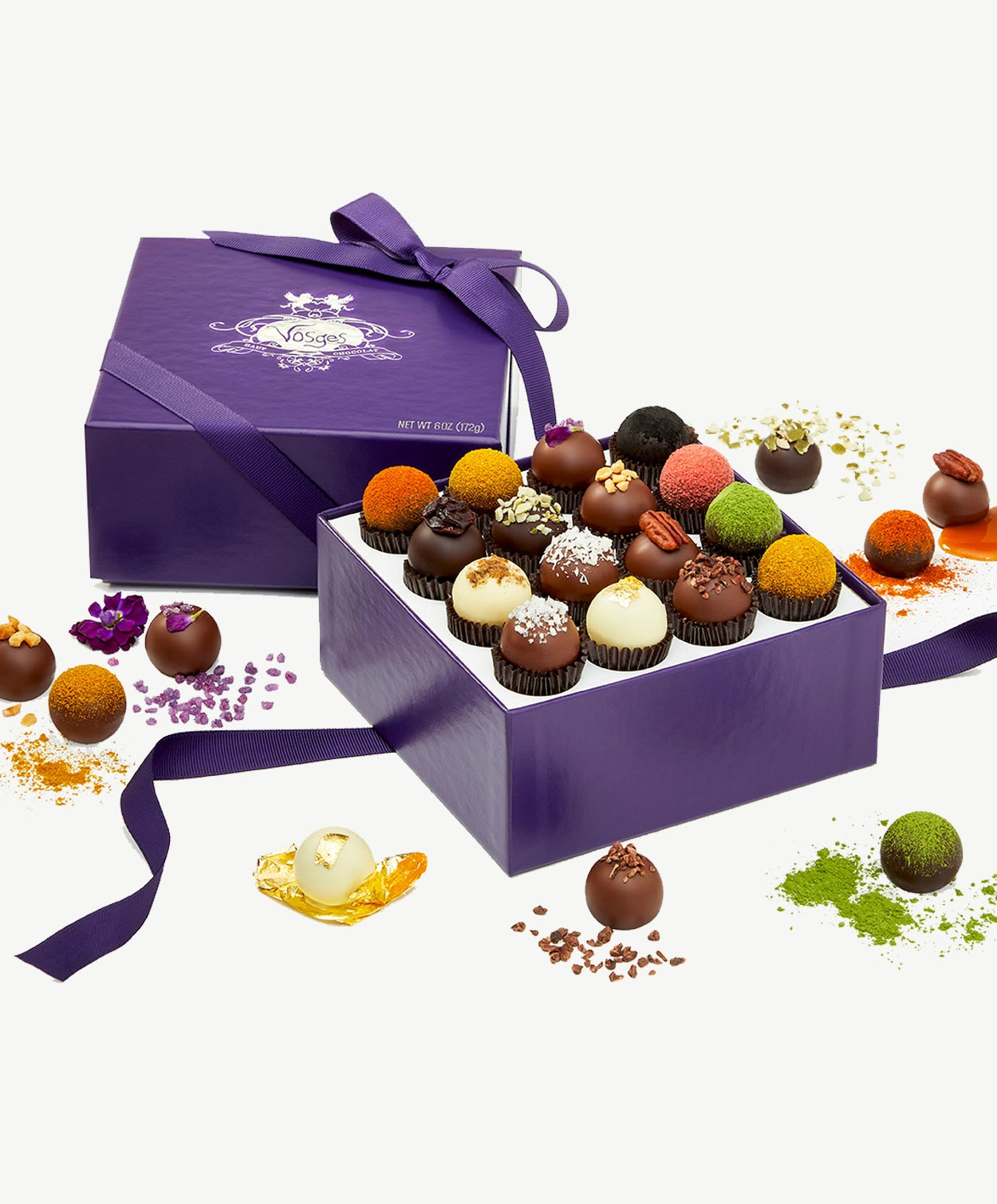 Open box of Vosges Haut-Chocolat surrounded by chocolate truffles and exotic ingredients on white background.