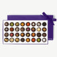 Large purple candy box of Vosges Haut-Chocolat sits horizontal displaying thirty two chocolate truffles adorned with colorful spices, chopped nuts and dried flowers tied with a purple ribbon bow on a grey background.