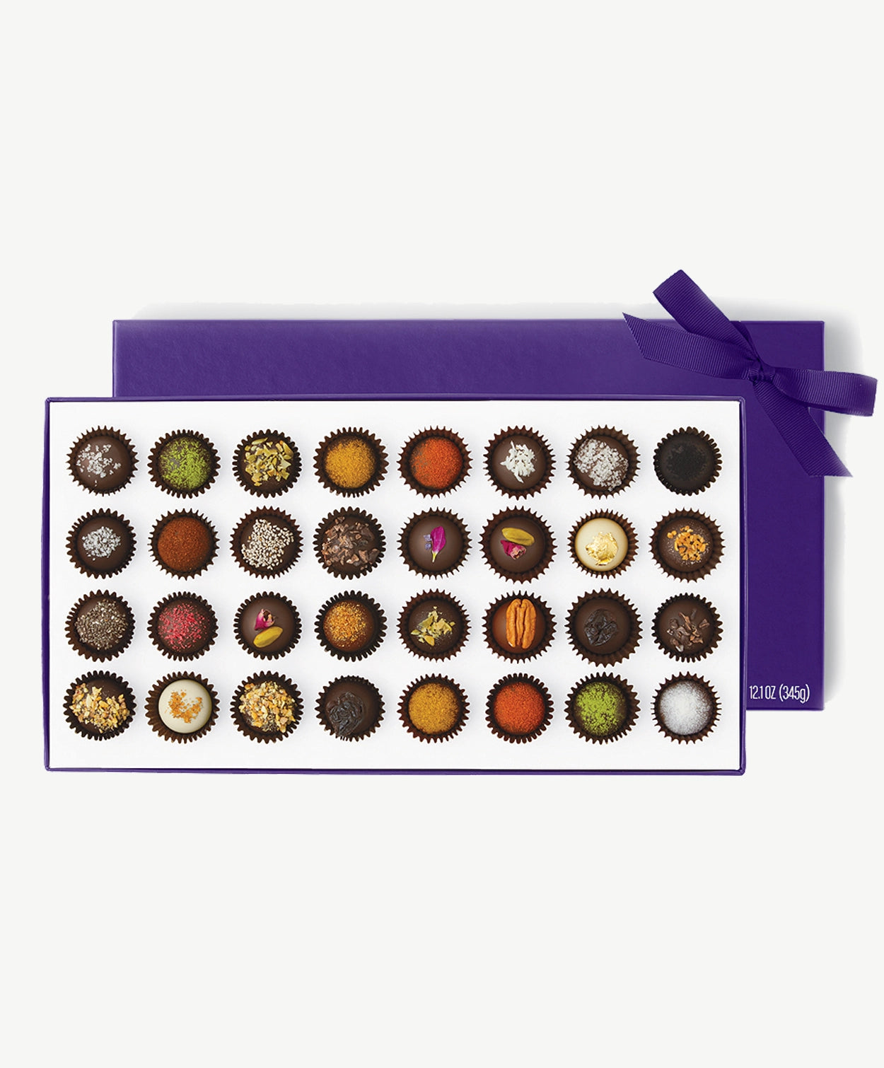 Large purple candy box of Vosges Haut-Chocolat sits horizontal displaying thirty two chocolate truffles adorned with colorful spices, chopped nuts and dried flowers tied with a purple ribbon bow on a grey background.