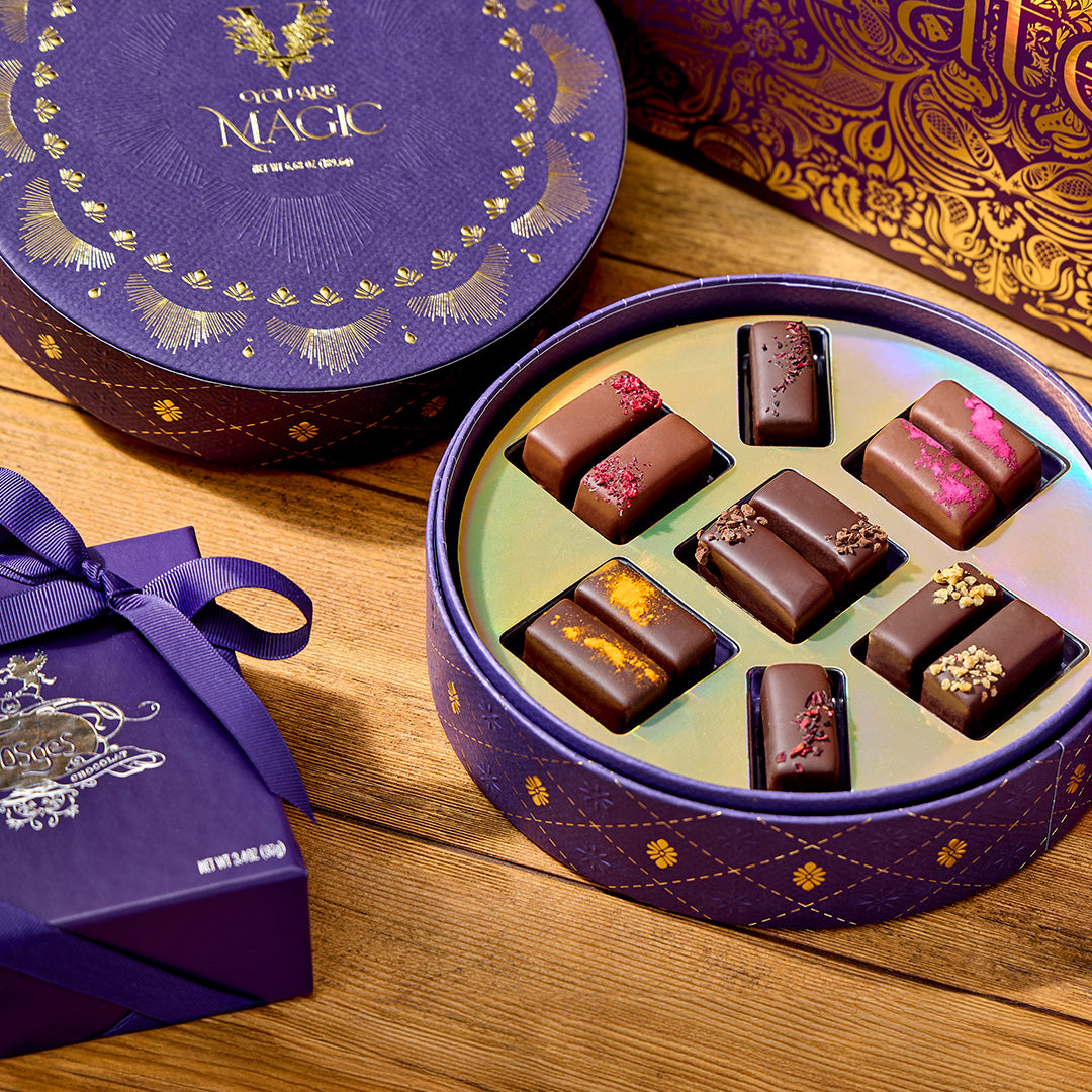 Purple round gift tin with gold patterns opened to reveal assorted chocolates in a gold tray on a wooden surface.