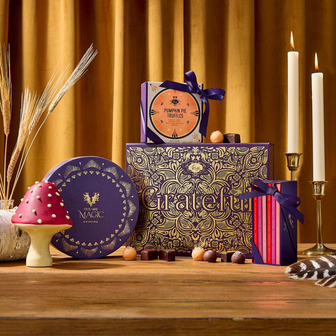 Festive The Giving Table gift boxes with intricate gold patterns, a purple round tin, chocolates, and pumpkins on a wooden table.