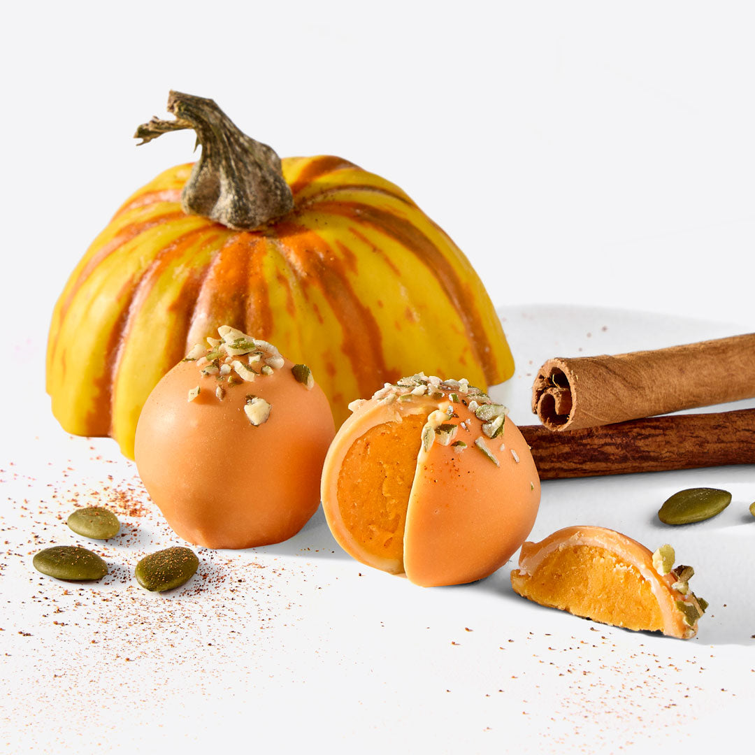 Orange coated Pumpkin Pie Truffles with pistachio toppings beside a small pumpkin and cinnamon sticks on a white surface